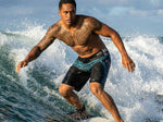 Surfer crouched on a breaking wave wearing HIC Kanaha black board shorts with teal Polynesian print