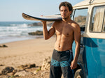 Man holding a surfboard on his shoulder beside a blue and cream camper van on the beach, wearing HIC Kanaha black and teal board shorts