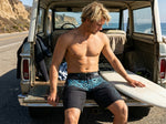 Man sitting on the open tailgate of a vintage truck beside a surfboard, wearing HIC Kanaha black board shorts with teal print, coastal road behind