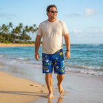 Man in a white T-shirt and sunglasses walking the shoreline in HIC Suck Em Up board shorts