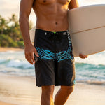 Close-up of HIC Kanaha 21-inch black board shorts with teal Polynesian print and drawstring waist, surfboard under the wearer's arm on the beach