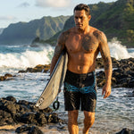 Man in HIC Kanaha 21-inch black board shorts with teal Polynesian print carrying a surfboard over wet black lava rocks, green mountains behind