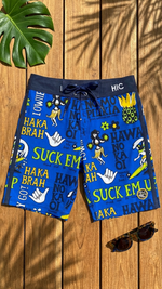 HIC Suck Em Up board shorts in blue laid flat on wood with sunglasses and a palm leaf